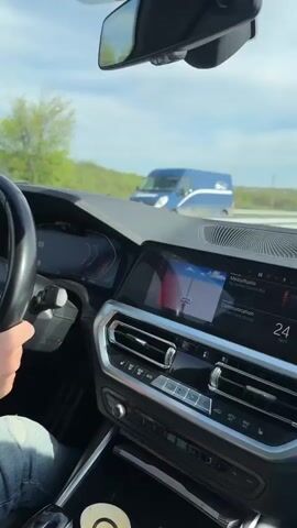 The media shows a first-person perspective from the driver's seat of a modern car, likely a BMW due to the visible emblem on the steering wheel. The driver's right hand, clad in ripped blue jeans, rests on the steering wheel, indicating the vehicle is in motion.

The interior features a sophisticated dashboard with digital displays. The instrument cluster shows various gauges and readouts, though specific numerical values are not clearly legible. To the right, a large infotainment screen displays a navigation map, with graphical elements suggesting a route. Below this screen are chrome-accented air vents and a control panel with buttons for climate and media, all part of the car's central console. A white cup is visible in a cupholder in the console area. The rearview mirror is also visible at the top of the frame.

Through the windshield, a bright, sunny daytime scene unfolds. The road stretches ahead, flanked by green fields or grassy verges under a clear blue sky dotted with scattered white clouds. The overall impression is of a calm drive on an open road.

The general setting suggests travel on a highway or rural road. The specific location context provided is Voyvodovo, Bulgaria, consistent with a driving scene. No specific legible text is visible on the screens or controls, apart from the BMW logo.