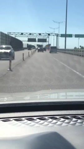 The video, shot from inside a moving vehicle through its windshield, captures a forward view of a multi-lane highway or bridge on a clear, sunny day. The sky is bright blue with no clouds.

In the foreground, the top of the vehicle's dashboard is visible. The road ahead is asphalt, showing some wear and tear, and is divided by short, orange and white striped poles or cones, suggesting ongoing roadwork or temporary lane division.

Several vehicles are seen driving in the distance. A large blue truck with a white trailer is on the left side of the road. Further down the road, closer to the center, are multiple passenger cars, including a dark-colored sedan. On the right, a white van is driving. All vehicles appear to be moving in the same direction.

Overhead, a complex metal gantry structure spans the width of the highway, supporting road signs and potentially other equipment. A prominent green road sign is visible on the right side of this structure, displaying white text. The main text reads "КРОНШТАДТ 14" (Kronstadt 14), indicating the distance to the city of Kronstadt. Below it, a partially visible English translation starts with "KRONSH". Tall lamp posts are also visible along the right side of the road, extending into the distance. To the left, a tall sound barrier runs parallel to the highway.