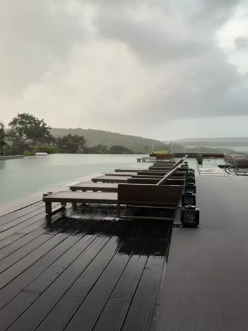The video shows an outdoor swimming pool on a cloudy, rainy day. In the foreground, a wooden deck with lounge chairs is visible, wet from the rain. The infinity pool stretches out behind the deck, with lush green trees and hills in the background. The sky is overcast with a hint of sunlight breaking through. Several lounge chairs are placed around the pool area. There are also some decorative elements near the pool, including a statue and some floating objects. The overall atmosphere is tranquil and somewhat moody due to the weather.