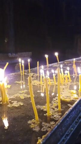 The video shows a close-up of several lit yellow candles floating in a shallow body of water. The candles are of various heights and some are partially submerged. Wax has melted and formed puddles around the base of many candles. The background is dark, suggesting an indoor environment, possibly a church or a place of worship. The camera is focused on the candles, with a shallow depth of field. The lighting is dim, primarily coming from the flames of the candles.