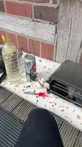 The video shows a patio or deck area next to a brick building. In the foreground, there is a wooden deck with a gray mat on the ground. To the left, there is a folding black chair and a roll of paper towels. On a white surface, there is a black pot with a lid, a bottle of orange liquid, and a large clear bottle of yellow liquid. There are also some tongs. To the right, there is a can of beer and some garlic. In the background, there is a green lawn, bushes, trees, and houses. In one shot, a woman is sitting on a chair with a stroller, holding a plate of food and a bag of chips. She is smiling and touching her mouth. The video appears to be filmed on a sunny day.