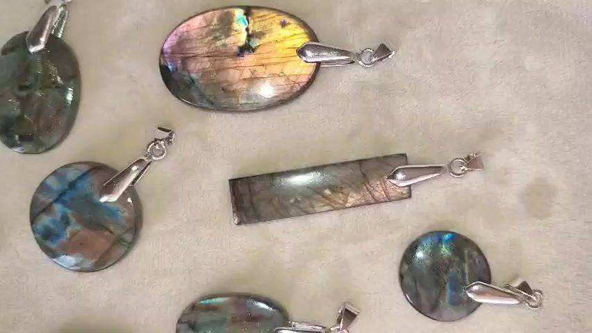 The video shows an overhead shot of several labradorite pendants laid out on a soft, textured surface, possibly fabric or felt. The pendants are of various shapes, including oval, round, and rectangular. Each pendant features a metal bail for attaching to a chain. The labradorite stones display a characteristic iridescence with flashes of blue, green, yellow, and pink light, particularly noticeable on the oval and round pendants. The lighting highlights the shimmering qualities of the stones.