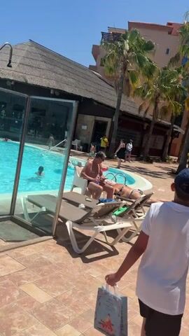 The video begins showing a portion of a swimming pool area with tiled flooring and lounge chairs. Several people are visible, including a man sitting on a lounge chair near the pool, a woman lying on a lounge chair, and a child walking away from the camera. The pool appears to be blue and people are in the water. Palm trees and a building with a thatched roof are also visible. The camera then pans up to the sky, showing a clear blue sky with a commercial airplane flying in the distance.