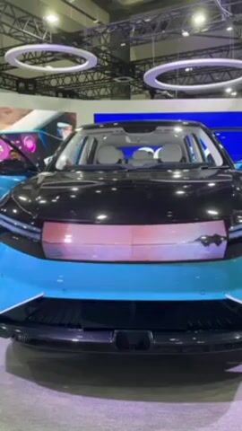 The video shows a stationary, light-blue and black car on display at what appears to be a convention center or showroom. The car has a screen embedded in the front grill. The car doors are open. The background includes overhead lighting fixtures in the shape of rings.