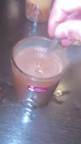 The video shows a person stirring a drink in a glass with a spoon. The drink appears to be chocolate-colored, and the glass has a red logo that reads “RED CHERRY.” There is steam rising from the drink. The scene is set at a table in what looks like a café or restaurant. Another drink, appearing to be a layered coffee beverage with milk or foam on top, is visible in the background. A man with a beard is sitting at the table. There are other people in the background, seated and possibly relaxing in the establishment. The table is dark-colored.