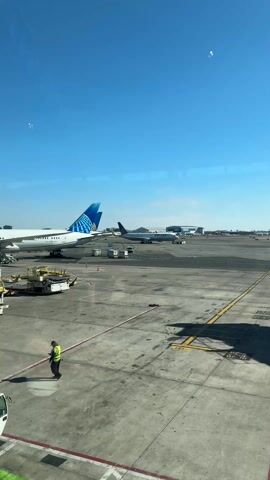 The video shows an airport tarmac on a clear day. A United Airlines plane is in the foreground, being towed by a vehicle. The plane is white with blue and gold accents on its tail. There are several pieces of equipment and a small white van near the plane. In the background, there is another United Airlines plane and an airport building. A person wearing a yellow vest walks across the foreground of the video. The view is from inside an airport terminal, looking out through a large window.