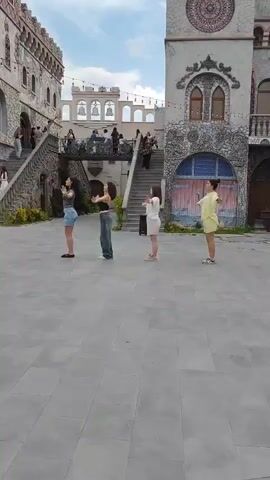 Four young women are dancing in a courtyard. The woman on the far left is wearing a gray tank top and denim shorts. The second woman from the left is wearing a black tank top and baggy jeans. The third woman from the left is wearing a white t-shirt and light-colored shorts. The woman on the far right is wearing a yellow dress. They are all facing the same direction and appear to be performing synchronized choreography. The courtyard has a stone floor and is surrounded by buildings that resemble a castle, with stone walls, arched windows, and a tower with a circular decorative element. There are people visible in the background on balconies and staircases. The sky is visible above, with some clouds.