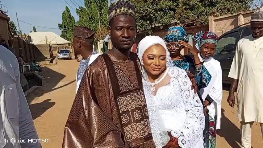 A wedding in progress. A couple, the groom in a brown traditional robe and matching cap, and the bride in a white lace dress and hijab, stand close together smiling for the camera.  They are surrounded by other people, some in traditional clothing. A black SUV is parked nearby. The location appears to be a residential area, with buildings and foliage visible in the background.