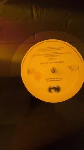 A record player is shown from above. The turntable is spinning, and a yellow record is on it. The record is black aside from the yellow center. The record center has black text and a logo. The environment appears dimly lit.
