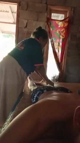 The video shows a man lying on his stomach on a massage table, receiving a leg massage from an older woman. The woman is wearing a dark blue T-shirt with some writing on the back and a long tan skirt. She has her hair pulled back in a bun and is using her hands to massage the man's legs. The man is wearing blue and white patterned shorts. The massage is taking place in a room with brick walls and two windows. A colorful piece of fabric is hanging in front of one of the windows. There is some sunlight streaming into the room.