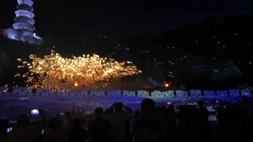 The video shows a night scene with a pagoda in the background on top of a hill, and a crowd of people in the foreground watching a fireworks display. The fireworks are golden and create a dazzling effect. The crowd is holding up their phones, presumably to capture the spectacle. There is a stone wall with battlements at the center. The surrounding environment is dark and hilly.
