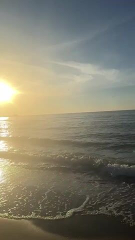 A video shows a beach at sunrise or sunset. The sun is low on the horizon, casting a warm glow over the water and sky. Gentle waves lap at the sandy shore, creating a peaceful and serene atmosphere. The sky is a gradient of blue and yellow, with a few wispy clouds. The camera is positioned on the beach, looking out towards the ocean.