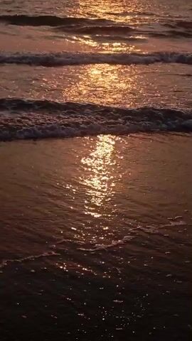 The video captures a serene ocean scene during sunset at Arambol, India. The setting sun, a bright golden orb, is visible near the horizon, casting a warm, shimmering reflection across the water. The sky above is a soft gradient of orange and pink.

Gentle waves roll towards the shore, breaking into white foam at the edges. The water closest to the camera is a dark, almost reddish-brown, reflecting the golden light of the sun in long, broken streaks that dance with the movement of the waves. The middle ground shows more defined wave patterns, each catching the sunlight and creating a sparkling effect.

There are no people or specific objects visible in the frame, focusing entirely on the natural beauty of the sunset over the sea. The atmosphere appears calm and tranquil. No text is visible in the video.