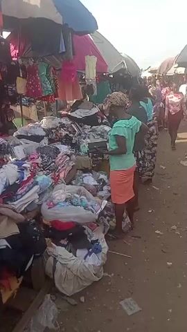 The video captures a bustling outdoor market scene in Umuahia, Nigeria. Numerous people, predominantly women, are seen navigating through narrow pathways lined with makeshift stalls. Large piles of clothing, ranging from dresses to shirts and other garments, are displayed on tables, tarpaulins on the ground, and hanging from racks under various colorful umbrellas and canopies providing shade.

Shoppers are actively browsing the merchandise, sifting through the piles of clothes, and occasionally picking up items to inspect them more closely. Vendors are present, attending to customers, although their faces are not always clearly visible. The atmosphere appears lively and busy, characteristic of a street market. The ground is unpaved, appearing like dirt or compacted earth. The time of day is clearly daytime, with bright natural light overhead. No specific text or signs are clearly legible within the frame.