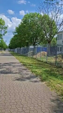 The video shows a paved walkway with a textured surface, bordered by grass and trees. A chain-link fence runs alongside the walkway, separating it from an area with some construction materials and vegetation. The sky is blue with scattered clouds, and sunlight casts shadows on the walkway. The scene appears to be an outdoor environment, possibly a park or a street with a pedestrian path. There are no people visible in the video, and the focus is on the landscape and the walkway.