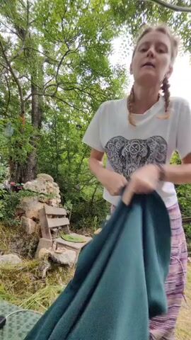 A young woman with braided blonde hair, wearing a white t-shirt with an elephant graphic and striped pants, is outside in a wooded area. She is in the process of putting on a dark green hooded sweatshirt. The setting appears to be a somewhat overgrown yard with a stone wall, a wooden bench, and scattered building materials. Trees and lush green foliage dominate the background. The lighting suggests it is daytime, likely overcast due to the diffused light. The woman's expression is neutral as she focuses on the task of dressing. There is no visible text in the frame.