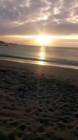 The video shows a beach at sunset. The sun is low on the horizon, shining brightly and casting a golden reflection on the water. The sky is filled with clouds, some of which are lit up by the sun. The sand is dark and wet, and there are small waves lapping at the shore. In the distance, a land formation can be seen.