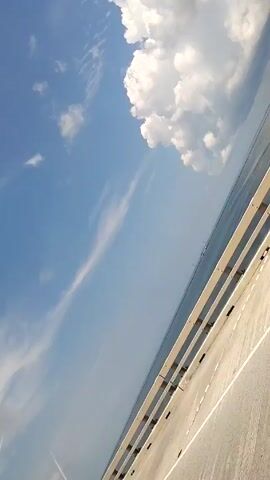 The video shows a view from a car driving on a long bridge. The bridge has a light gray surface and a metal guardrail. To the right of the bridge is water. The sky is blue with large, fluffy white clouds.  The perspective is slightly tilted upward and to the right, giving the impression of the sky dominating the view.  A faint jet stream is visible in the lower left corner. Light glares are visible on the car window.