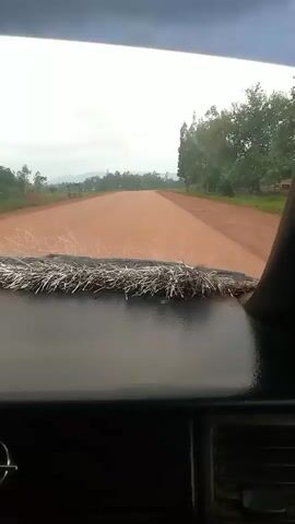 The video shows a view from inside a vehicle looking out the windshield at a dirt road ahead. The road is reddish-brown and appears to be unpaved. There are trees on either side of the road, and some distant hills or mountains can be seen in the background. The sky is overcast and appears to be a light gray. The dashboard of the vehicle is visible in the foreground, with some decorative material on top of it.