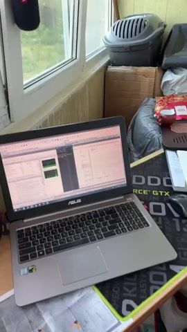 The video shows a cluttered desk area on a balcony or enclosed porch. A laptop is open on the desk, displaying what appears to be some sort of software interface. To the right of the laptop are various items including a computer mouse, a notebook, and what looks like a gaming mousepad. Further to the right, on a windowsill, is a collection of camera equipment, including a DSLR camera with a lens attached, several battery chargers, and small LED lights. The background shows a window with curtains and some boxes and other miscellaneous items stacked behind the desk. The scene appears to be an improvised workspace or hobby area.