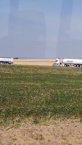 The video shows a truck with a white tank and the word "UNIVAR" printed on it driving on a road in a rural area. There is a field of green grass and some brown vegetation in the foreground. In the background, there is a field of yellow crops and a blue sky with some clouds. Another truck is visible in the distance on the left side of the frame. The camera appears to be moving, likely from a vehicle, as the landscape is in motion.