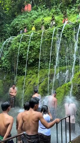 A group of people stand in shallow water at the base of a moss-covered rock face. Numerous spouts of water cascade down from above, some directed at the individuals. The water appears to be falling from pipes running horizontally across the cliff face. Several men are visible, some partially submerged, others standing and facing the waterfalls. The environment is lush and green, with dense foliage growing on the rock face. In the foreground, several individuals are facing away from the camera, looking towards the waterfalls. Some are wearing sarongs, and others appear to be in bathing attire. A railing is visible in the lower right corner of the frame.