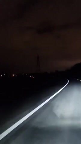 The video shows a first-person perspective from a vehicle driving on a dark, winding road at night. The only visible light source is the vehicle's headlights, which illuminate the white line marking the edge of the road. In the distance, there is a tall transmission tower visible against the dark, cloudy sky. There are faint lights from what appears to be a distant settlement on the left. The road curves to the right.