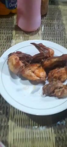 A plate of fried chicken wings is shown on a table. The chicken is golden brown and appears to be seasoned. There are several pieces of chicken on the plate. In the background, a pink cup or container is visible, along with a blurry bottle. The table surface appears to be made of woven material. The lighting is dim.