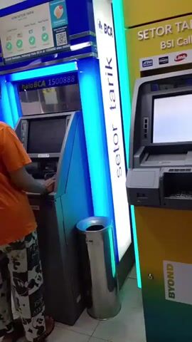 A woman in an orange t-shirt and patterned pants is at an ATM. To her left, another ATM has a person in an orange shirt using it. To her right, another ATM is visible. A metal trash can is between the two ATMs. The ATMs are in a brightly lit indoor space with tiled floors.