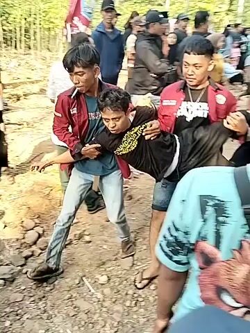 A group of young men are carrying another young man, who appears to be injured or unconscious, through an outdoor area. The scene is somewhat chaotic, with other people observing and some appearing to be involved in assisting. The environment seems to be a rural or semi-rural setting, with dirt ground and some simple structures visible. The men carrying the injured person are wearing matching red shirts with white lettering on the back, suggesting they are part of a group or organization. The lighting suggests it is daytime. The people are dressed in casual clothing.