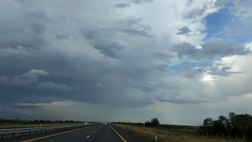 The video is a first-person perspective from inside a vehicle traveling on a highway. The sky is filled with dramatic, dark storm clouds, with patches of blue sky visible in the distance. The road ahead is empty except for a few distant vehicles. The landscape on either side of the highway is dry and sparsely vegetated with some trees and bushes. The overall atmosphere is moody and suggests an approaching storm.