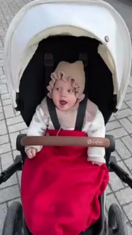 A baby is sitting in a stroller. The baby is wearing a beige knit hat with ruffles around the edge, a beige and white patterned sweater, and a beige scarf. The baby is buckled into the stroller with black straps. The baby is covered with a red blanket. The stroller is black and white, with a brown handle labeled "cybex". The stroller is on a paved surface. The baby is looking around.