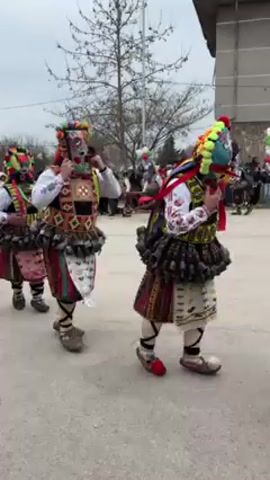 A group of people in colorful costumes and masks are walking in a procession on a paved road. The costumes consist of embroidered fabrics, bells, and decorative headwear.  The road appears to be in a village, and there are other people watching the procession. Some buildings and bare trees are visible in the background, under an overcast sky.