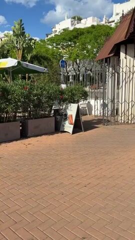 The video shows a street scene in a sunny location. First, it focuses on an A-frame sign advertising "Chocolate con Churros" for 4.50 at Cafe Bar Europa. The sign features a picture of the chocolate and churros. Next, it pans up to show tables and umbrellas outside a cafe. The scene includes green shrubbery in planters. Finally, the video pans across a brick sidewalk towards a green space with trees in the background.