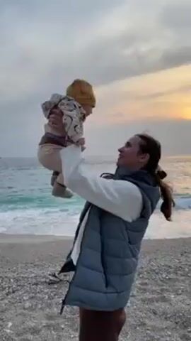 A woman and baby are on a beach. The woman holds the baby up in the air. The woman is wearing a white sweater, a gray vest, and brown pants. The baby is wearing a yellow hat, a patterned jacket, and tan pants. The beach is rocky. The water is blue with white waves. The sky is gray and the sun is setting on the horizon.