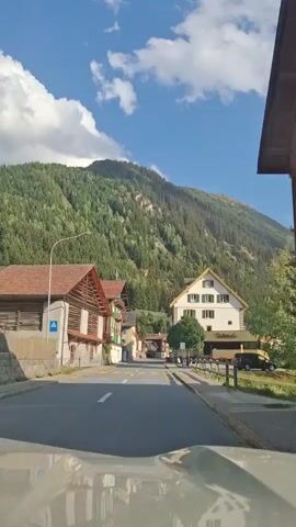 The video shows a forward-facing view from inside a vehicle driving down a street in a village nestled at the base of a forested mountain. The sky is blue with scattered white clouds. On the left side of the street, there are traditional wooden buildings with red roofs and some with green shutters. One building has "POST" visible on its facade. On the right side of the street, there are more buildings, including a larger one with a yellow roof and a sign that appears to say "HOTEL RISTORANTE". A tree stands in front of this building. The street is paved and has yellow crosswalk markings. The vehicle is moving forward at a moderate pace.