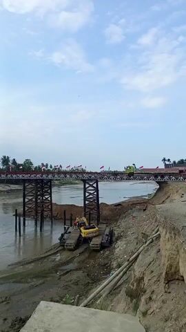 The video shows a view of a river with a bridge stretching across it. The bridge is adorned with numerous Indonesian flags. A green truck travels across the bridge from right to left. On the riverbank in the foreground, a yellow excavator sits on a floating platform. The riverbank appears to be a sandy and muddy environment. The sky is blue with scattered clouds.