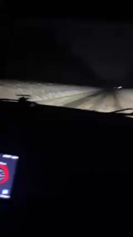 The video shows the interior of a car driving at night, with a snowy road visible through the windshield. The dashboard's touchscreen display is lit up with a red and black interface. A hand briefly interacts with the screen. The car's speedometer and other gauges are also visible. The road ahead is dark with a triangular warning sign illuminated on the roadside.