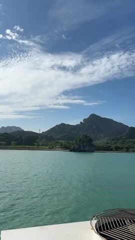 The video shows a view from a boat on a calm, turquoise body of water. In the distance, a lush green coastline is visible, with rolling hills and mountains covered in dense vegetation. The sky is a clear blue with scattered white clouds. A small rocky island with some greenery on top sits in the foreground of the coastline. Part of the boat's railing or deck is visible in the lower right corner of the frame. The camera appears to be moving slightly, likely due to the gentle motion of the boat.