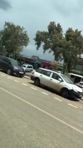 The video depicts a street scene with several vehicles parked on the side of the road. The primary focus is on a white station wagon with visible damage to its front end. Other vehicles, including a dark-colored car, a white van with yellow striping, and a black SUV, are also visible. In the background, there are trees, buildings with colorful facades, and people walking. The weather appears cloudy or overcast. The camera perspective seems to be from inside a moving vehicle, looking out at the scene.