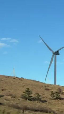 The video shows a wind turbine atop a brown, grassy hill under a bright blue sky with scattered white clouds. The turbine is gray and has three long blades. An overhead power line runs along the bottom of the hill. There are a few small trees scattered on the hill. The camera appears to be moving, possibly from a passing car.