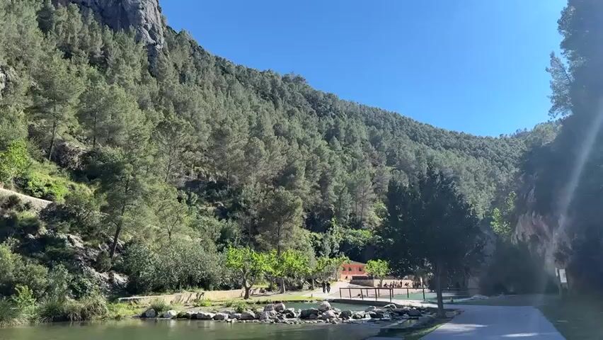 The video shows a mountainous landscape with a river in the foreground. The mountains are covered in dense green forest, with some rocky outcrops visible. A winding road runs along the side of the mountain. Below the mountain, there is a calm, green river with rocks along the bank. A small building with a red roof can be seen nestled among the trees near the river. The sky is clear and blue.