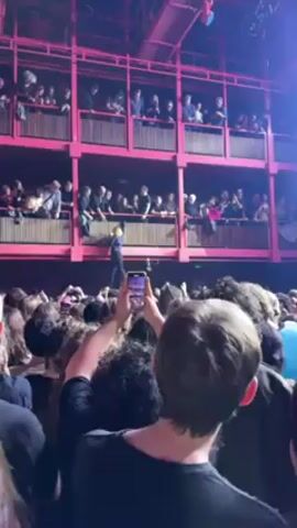 A crowd of people are standing and raising their arms and phones at a concert venue. A man is standing on a balcony above, gesturing to the crowd. The balconies are part of a tiered structure painted in bright pink. Many people in the crowd are recording the scene with their phones. Some of the crowd members are smiling. The venue's interior appears dimly lit with stage lighting.
