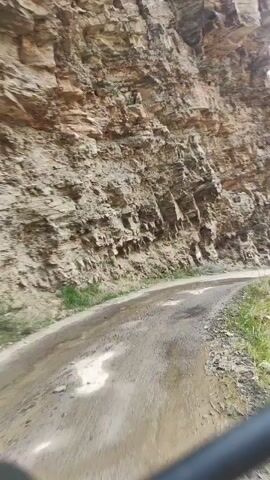 The video shows a POV shot of a dirt road winding alongside a massive rock face. The road appears wet in places, possibly from recent rain. The rock face is rough and layered, with signs of erosion. In the distance, a valley opens up with mountains on the other side. The sky is overcast with some clouds. The overall impression is a remote and rugged natural landscape.