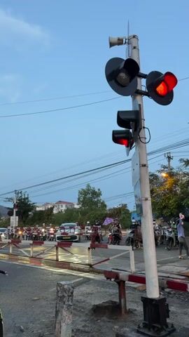 The video shows a train passing a railroad crossing in Vietnam. The crossing is protected by red flashing lights and a crossing gate. Numerous motorbikes and a car are waiting on the road for the train to pass. A man on a motorbike waits on the far side of the crossing. The train is made up of passenger cars followed by freight cars. The sky is clear and blue.