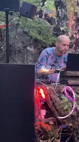 The video shows two men operating DJ equipment outdoors in a rocky, wooded area. The man on the left, bald with tattoos on his neck, is wearing a colorful t-shirt and is looking at a laptop. The man on the right, wearing a black t-shirt and a baseball cap, is looking at a DJ mixer with "Pioneer" visible on it. There are speakers, cables, and neon lights set up around the DJ booth, which is constructed from wood and branches. The background features large rocks and trees with dappled sunlight.