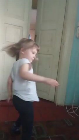 A young girl stands in a room. She wears a white shirt with a graphic on the front and dark-colored pants. She has light brown hair. Behind her is a slightly ajar white door that leads to another room, the interior of which is partially visible. The room she stands in has a patterned rug on the floor. She makes a few gestures with her hands. There is also a white electrical cord visible on the floor and a dark garment on a piece of furniture near the door.