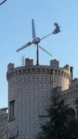 The video shows a homemade wind turbine on top of a brick tower. The wind turbine has three blades and is attached to a pole. The tower is made of light-colored bricks and has a crenellated top. There is a window on the tower, and a tree branch is visible in the foreground. The sky is blue. The blades on the turbine are spinning at varying speeds, with one blade appearing to be broken.