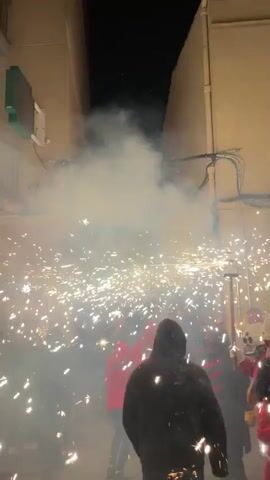 The video shows a nighttime outdoor scene, possibly a street or alleyway, with a group of people. The area is filled with a large amount of smoke and numerous bright sparks, suggesting the use of fireworks or pyrotechnics. Several individuals are visible, some wearing hooded garments and others in red outfits. One person in the foreground is wearing a white hat and a bandana around their neck, and is touching their hat. Another person is holding a tool with a circular object at the end, which appears to be a firework or a prop. The background consists of building walls and electrical wiring. The overall atmosphere is energetic and chaotic due to the pyrotechnics and the gathering of people.