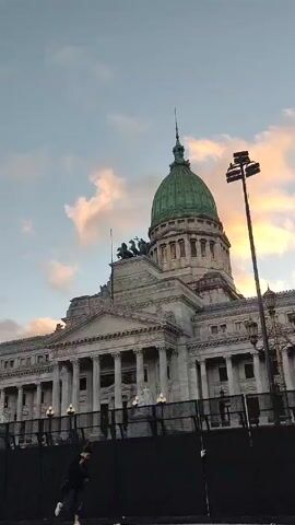 The video captures a view of the Palace of the Argentine National Congress in Buenos Aires, Argentina, under a sky painted with soft hues of dawn or dusk. The building is a large, imposing structure with neoclassical architecture, featuring a prominent green dome topped with a spire. The facade includes multiple columns, statues, and intricate detailing. A black barrier, seemingly made of connected fence panels, runs along the front of the building. A person dressed in dark clothing and light-colored sneakers walks slowly in front of this barrier.  Streetlights and a tall light pole are visible on either side of the building. The overall scene evokes a sense of grandeur and formality.